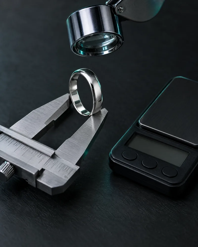 Illustration of a plain silver ring held in steel calipers under a jeweler's loupe, next to a pocket scale